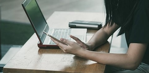 Person's hands using a smartphone next to an open laptop on a wooden desk.