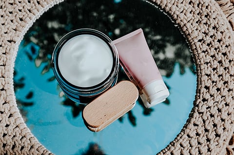 Open jar of white cream, pink tube, and wooden soap dish on a reflective glass table.