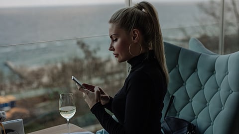 Woman sitting by a window near the sea, using her phone while having a drink.