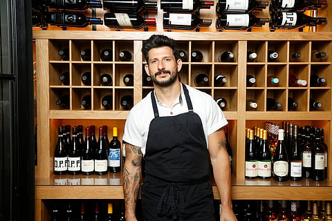 Chef Giorgio Rapicavoli stands before a floor-to-ceiling wine rack at Luca Osteria in a white shirt.