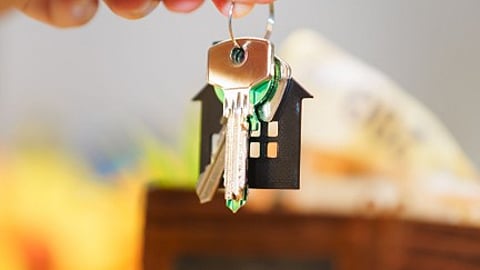Hand holding house keys above coins and a model home.