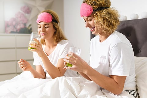 Two women in bed wearing sleep masks and holding drinks.