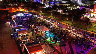 Aerial view of a crowded beachfront festival tent and stage at Lummus Park in Miami Beach.