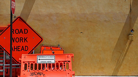 Road work signs and orange barriers leaning against a yellow wall.
