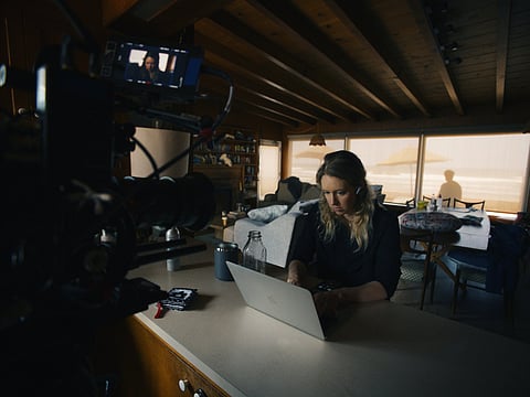 Elizabeth Holmes working on a laptop at a kitchen counter as a film camera records her