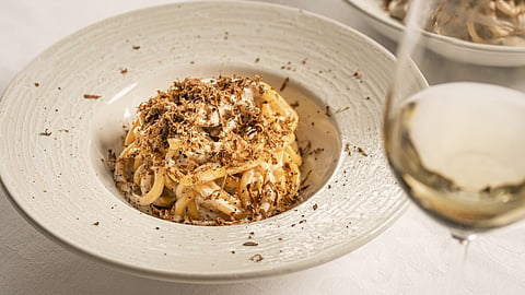 Bowl of cacio e pepe topped with shaved black truffle beside a glass of white wine
