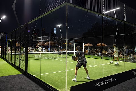 Two padel players rally on a green glass-walled court at night with spectators beyond