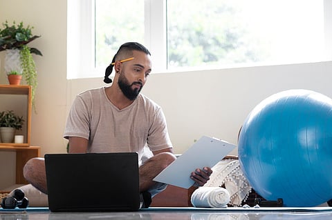 Fitness coach reading clipboard beside laptop and exercise ball.