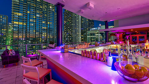 Rooftop bar bathed in pink light with high-rise towers in the background at dusk