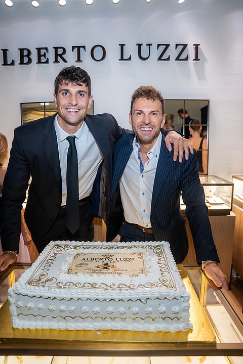 Two men in dark suits stand behind an anniversary cake in a jewelry boutique