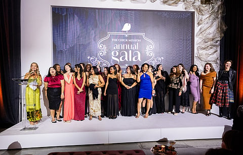 A group of women in evening wear stand on a stage before an Annual Gala backdrop