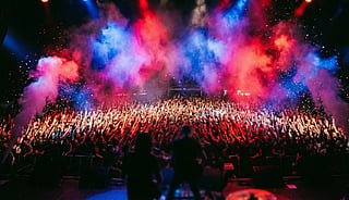 Concert crowd with raised hands beneath clouds of red, blue, and purple stage smoke