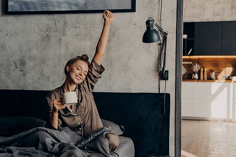 Sleepy relaxed lovely girl in satin pajama trying to wake up from night sleep, sitting in bed with closed eyes while holding her cup of coffee and stretching hand up, enjoying weekend at home