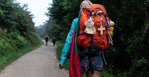 A woman walking a forested path on the Camino de Santiago with an orange backpack decorated with seashells.