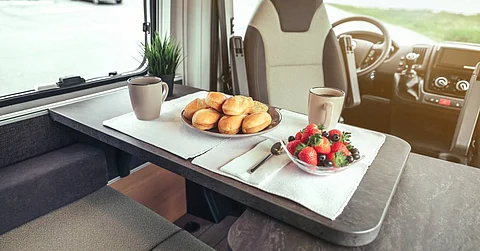 An RV parked during snack time with the table set featuring a bowl of fruit and a plate of bread options.