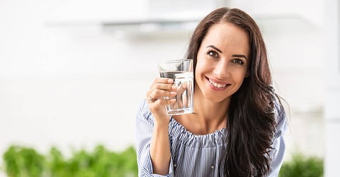 A woman stands in her kitchen, smiling while holding a glass of water. Plants sit on the counter behind her.