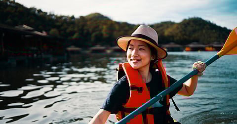 A woman in a life jacket and hat paddles through the water in a kayak. Several buildings are along the shore.