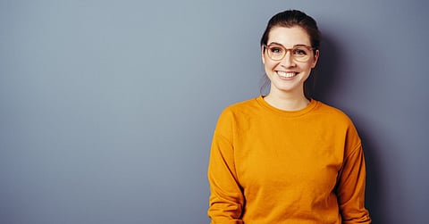 A young woman wears a yellow sweater and glasses with a brown frame. The background is a standard grey color.