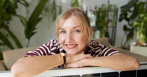 A happy woman sits in her home with her arms folded on a solar panel. She wears a red and white shirt.