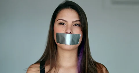 A woman with long, dark hair is in front of a dark grey background. She has a piece of duct tape over her mouth.