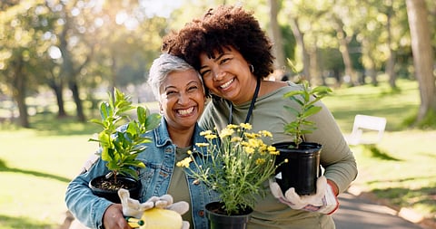 Two smiling women hold potted plants and gardening tools during a community volunteer event at a local park.