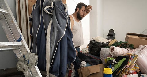 A man looking into a cluttered room with a serious look on his face. There are piles of items and boxes in the room.