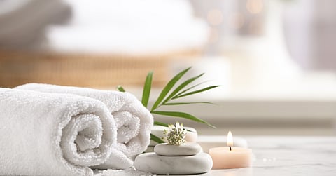 Two white, rolled-up towels sit next to a green leaf, two flat, white rocks, and a small lit candle on a white surface.