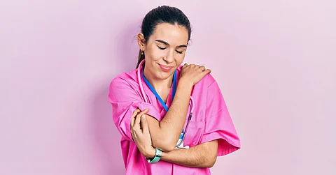 A young woman wearing a pink nurse uniform and stethoscope around her neck is smiling and hugging herself.