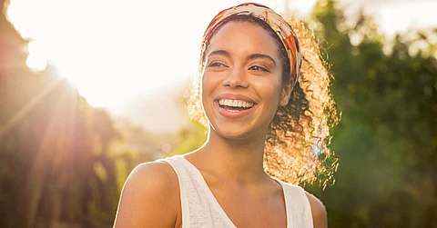 A smiling young woman wearing a bandanna over her curly hair standing in front of some trees with the sun in the background.