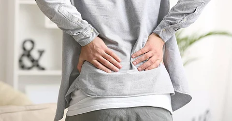 A young adult holding their lower back feeling chronic pain in a modern living room full of natural lighting.