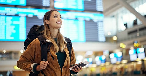 A woman smiles as she stands in an airport, wearing a backpack and holding her passport. Flight departures are behind her.