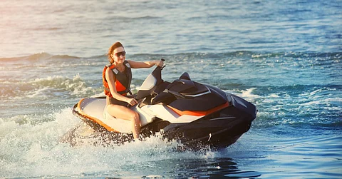A woman wearing sunglasses and a red and black floatation vest rides a jet ski through the water and makes splashes.