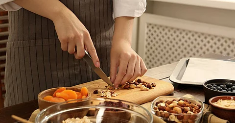 A person wearing a striped apron chops mixed nuts on a wooden cutting board, with bowls of dried apricots and other food nearby.