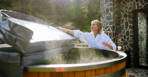 Woman wearing a white robe outdoors taking the plastic round cover from a hot tub while steam comes out.