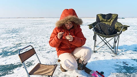 A woman wearing a warm coat sits on a chair on the ice as she prepares her bait. She has two other rods on the ground.