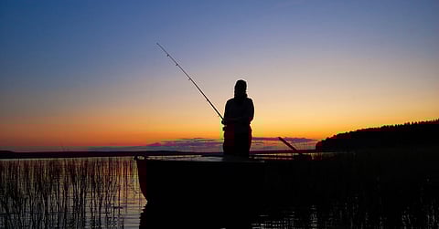 An angler holding a fishing rod while standing on a boat in the reeds as the sun sets in the background.