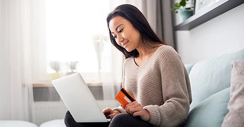 A young woman sitting cross-legged on her couch holds an orange credit card in one hand while using a laptop.