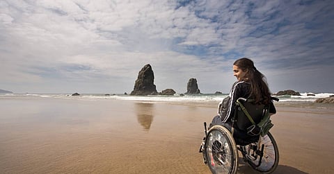 A young woman in a wheelchair on the beach at low tide with large rock structures coming out of the water.
