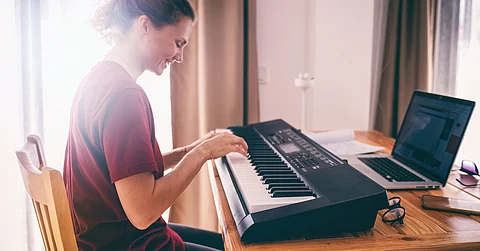 A woman sits at a table with an electric keyboard and an open laptop in front of her with her hands over the keys.