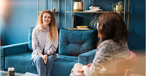 A woman smiling as she sits on a couch in her therapist's office. The sofa and the office walls are blue.