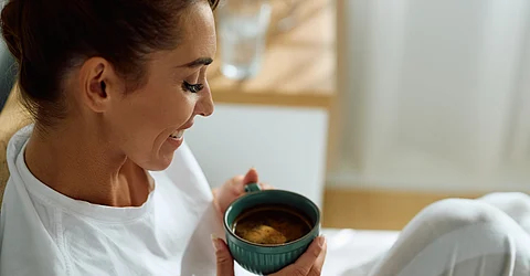A woman wearing a white shirt and pants holds a green mug full of brown liquid while sitting in bed.