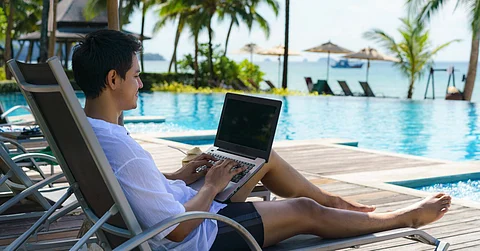 A man lays on a tan lounge chair next to a pool with his laptop on his lap. Palm trees appear in the background.