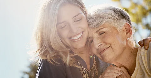 A young woman with long hair holding a senior woman close to her face with an arm wrapped around her back.