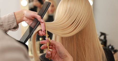 A person uses gold scissors to cut the hair of a person with blonde hair. They also hold a black comb in their fingers.