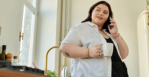 A woman with black hair standing in her kitchen and talking on a smartphone while holding a white coffee mug.