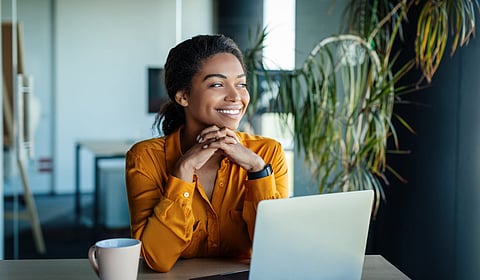 A smiling woman sitting at a desk with a laptop and coffee cup on it. A potted plant is visible behind her.