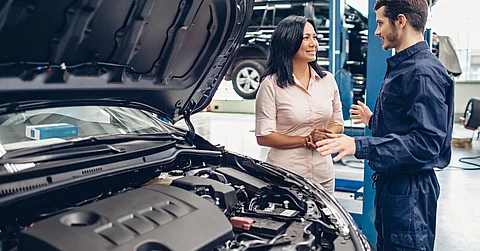 A woman talks to a mechanic in a blue jumpsuit at a car center. The hood on the vehicle next to them is open.