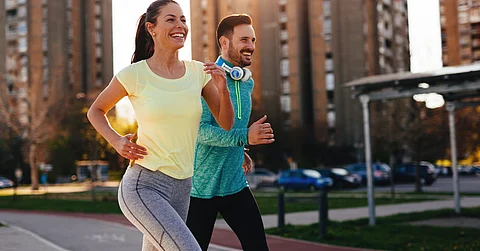 A woman and man wearing athletic clothes while running outdoors through a street with multiple buildings on a sunny day.