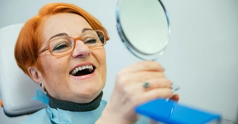 A red-haired senior woman with glasses sitting in a dentist chair while looking at her teeth in a handheld mirror.