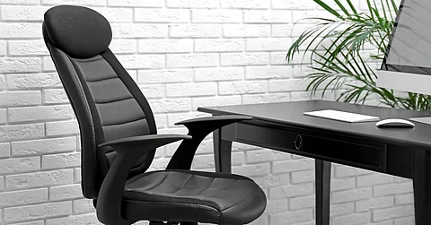 A black office swivel chair with a padded headrest and cushions sits in front of a desk with a computer.
